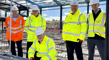 Five men, in Hi Vis jackets and hard hats stand on a building site underneath a steel frame of a building