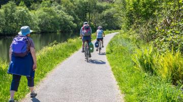 A woman walks along a path next to a river with two cyclists ahead of her