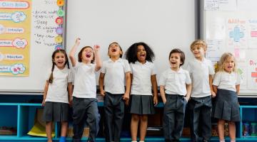 Image shows a line of primary school children in uniform standing in front of a whiteboard laughing 