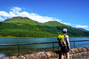 Walker with yellow rucksack walking alongside a wall with metal railing beside a loch looking across to green mountains