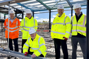 Five men, in Hi Vis jackets and hard hats stand on a building site underneath a steel frame of a building