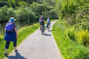 A woman walks along a path next to a river with two cyclists ahead of her