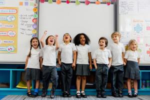 Image shows a line of primary school children in uniform standing in front of a whiteboard laughing 